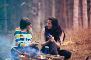 Two women sitting in the forest in front of a fire having a conversation