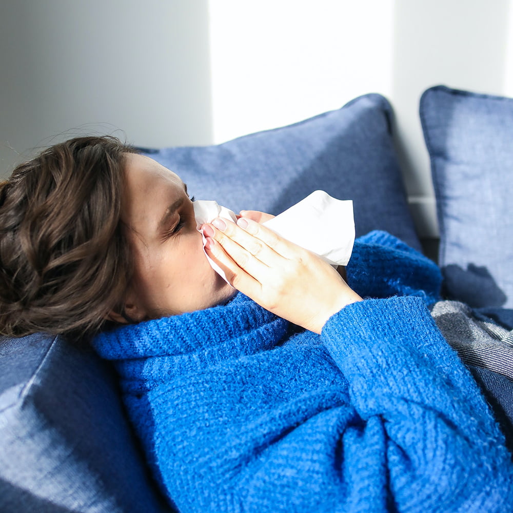 Brown haired woman in blue jumper sneezing into tissue