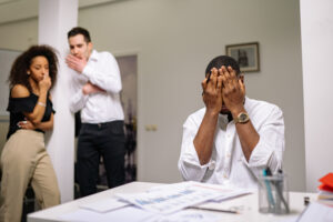 Frustrated employee covering eyes with hands while two other employees look at him, whispering to each other