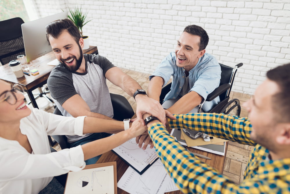 Office employees sitting around desk, smiling, putting their hands together