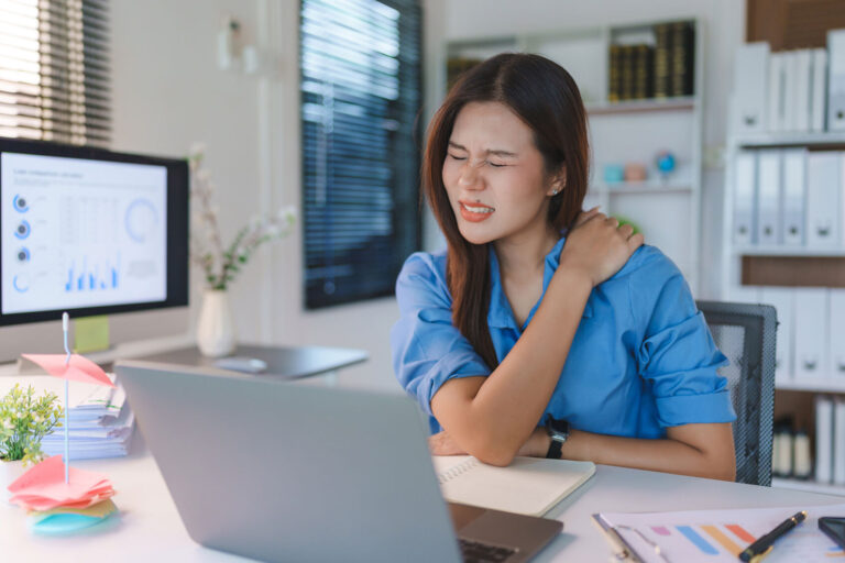 A woman sitting at office desk holding her shoulder in pain.