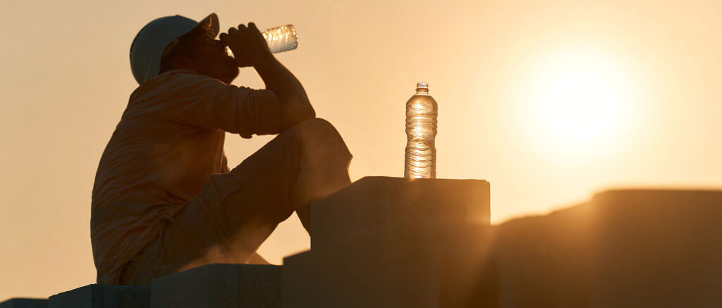 A worker sitting near sun with his water bottles.